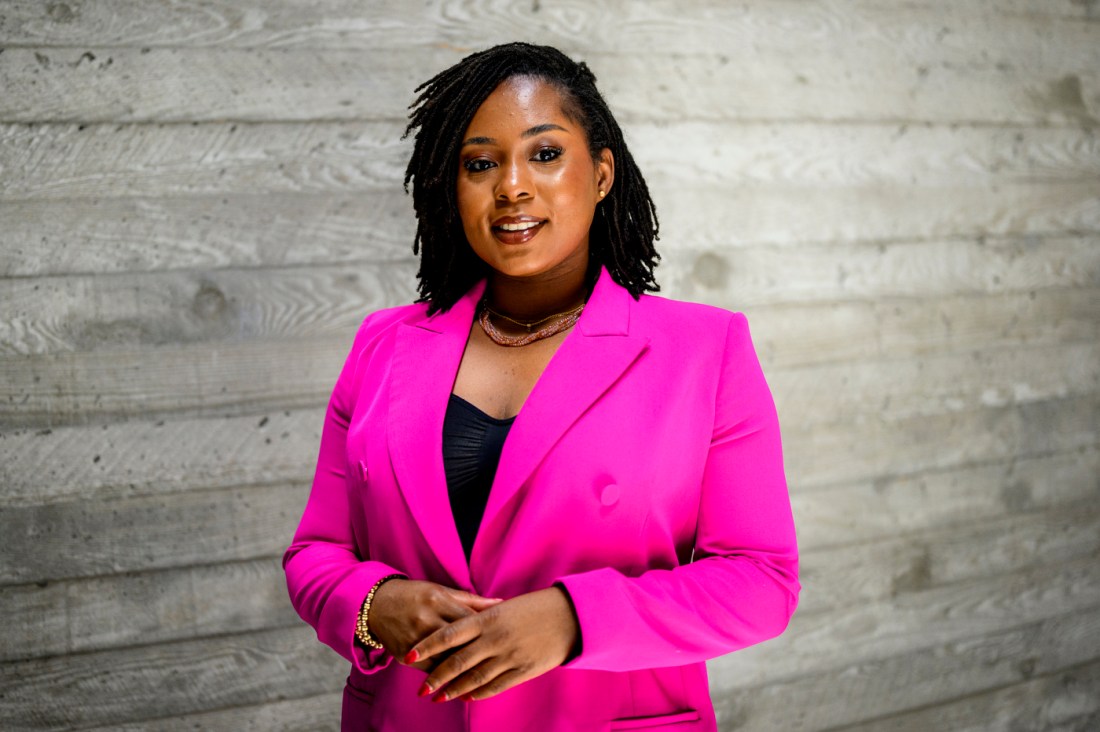 Portrait of Nita Akoh wearing a pink blazer in front of a wooden panel background.