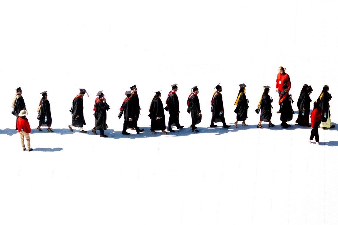 Graduates in caps and gowns walk in a procession across a bright surface.