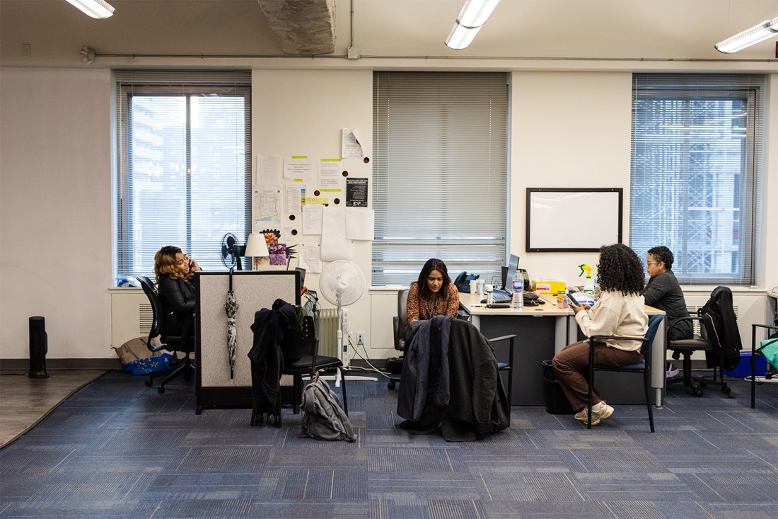 Students sit around a table and at a desk in a classroom. Some are on the phone and some are working on their laptops.