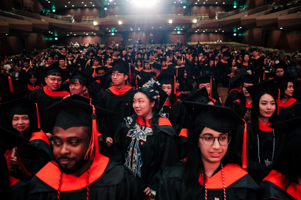 Rows of Northeastern graduates wearing black caps and gowns with red accents seated inside of Benaroya Hall for the Seattle Campus Commencement Ceremony.