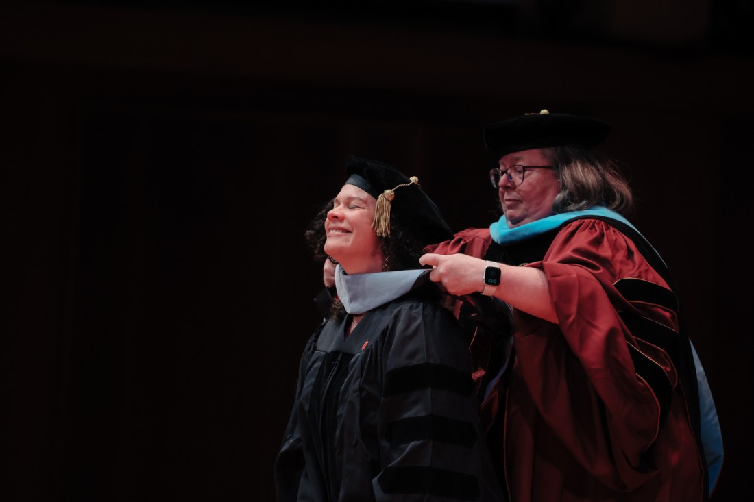 A faculty member in maroon academic regalia with blue accents places a doctoral hood around a smiling graduates neck.