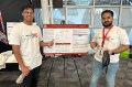 wo people stand beside a display board and smile for a photo at a presentation event.