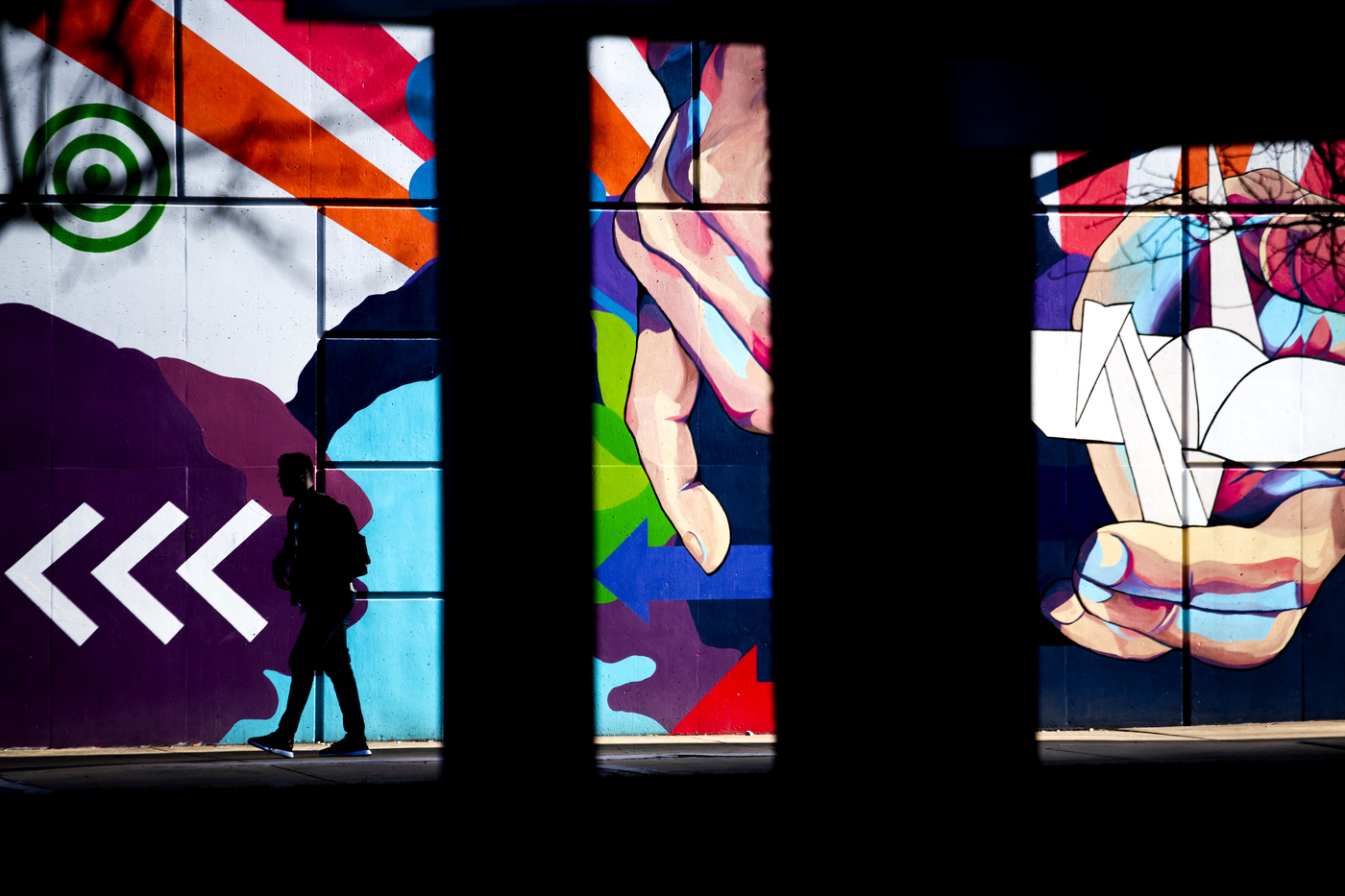 Pieces of two murals, separated by columns. Both have bright vivid colors and stripes, with other graphical elements such as arrows. On the right, a hand holding an origami crane draws focus.