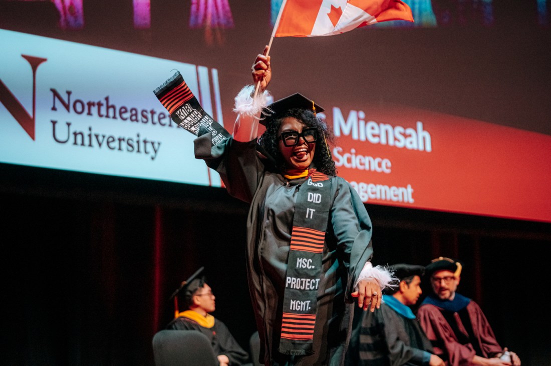 A graduate wearing black commencement reglia walking across stage waving a Canadian flag and sticking their tongue out.