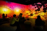 People sit in a dark room surrounded by colorful, abstract visuals projected on the walls.