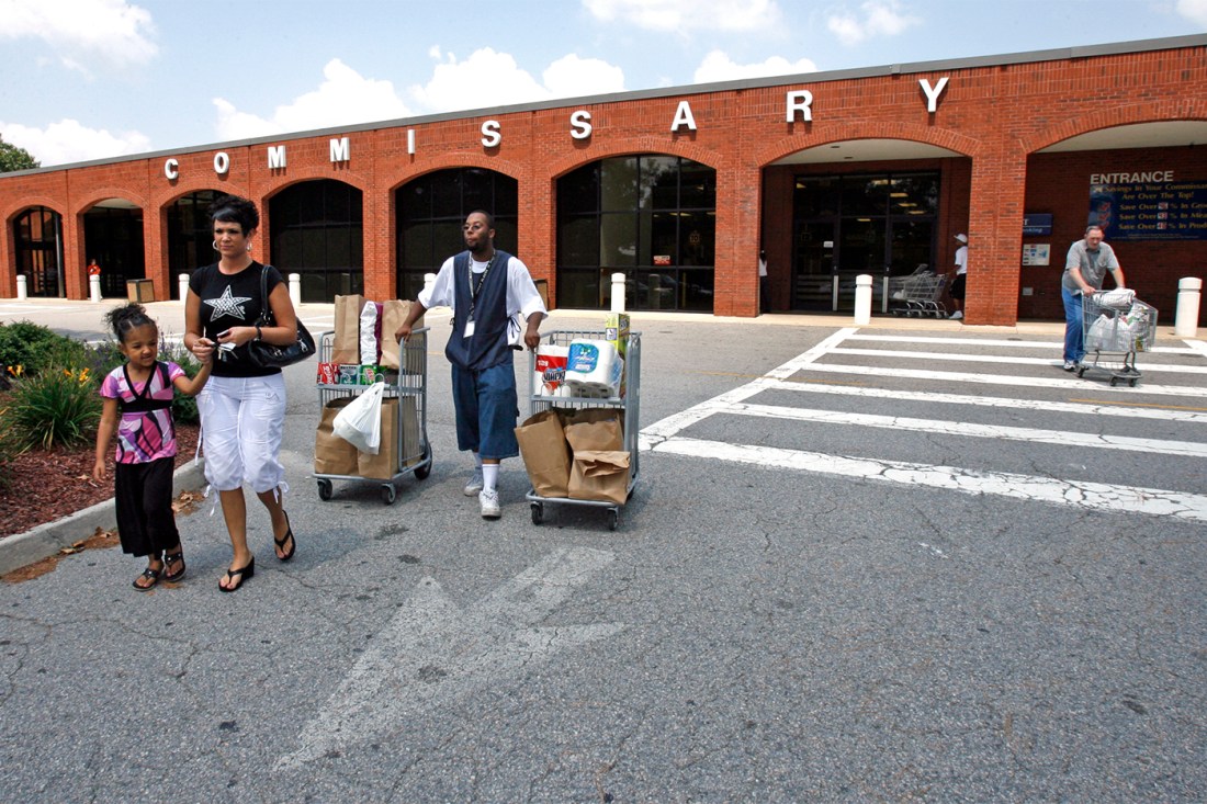 A group of people walking, some moving mobile carts, outside a brick building with "COMMISSARY" spelled out in large white letters across the top