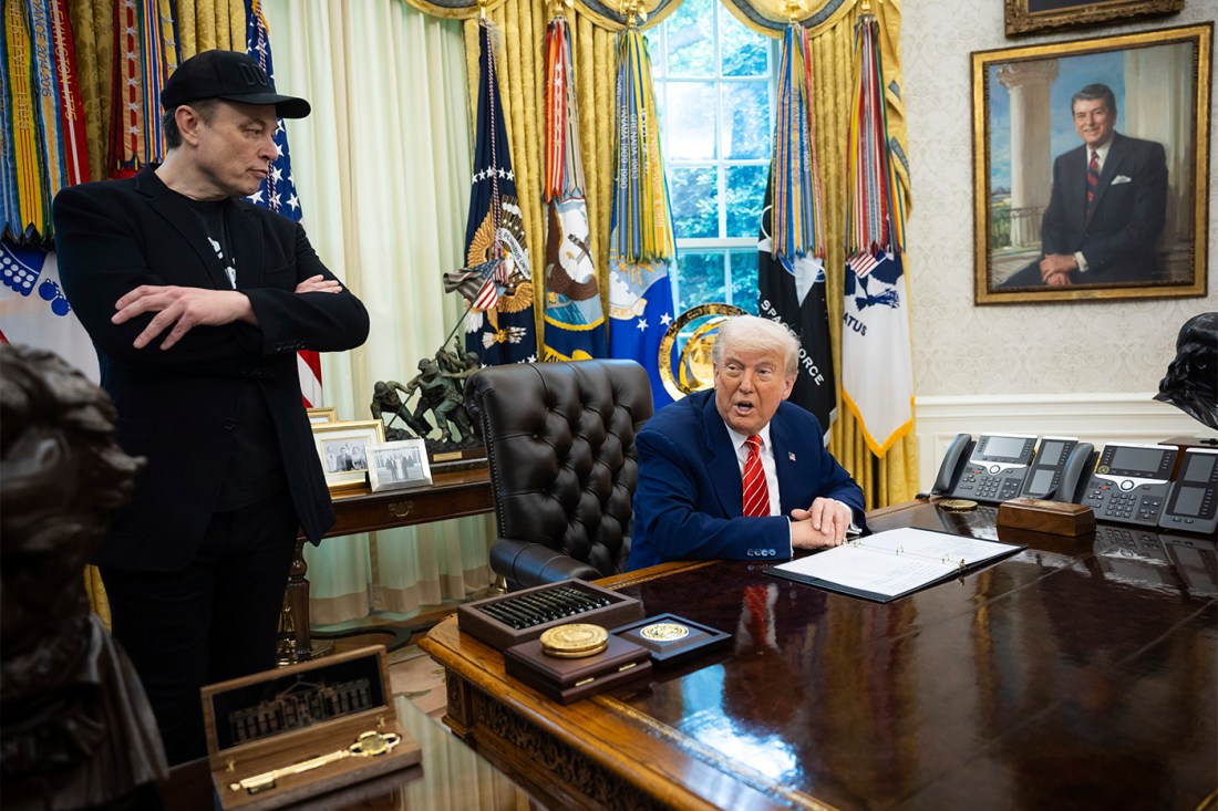 Donald Trump sitting at a desk in the oval office, in the middle of saying something. Next to him, Elon Musk is standing wearing a baseball cap with his arms folded across his chest.