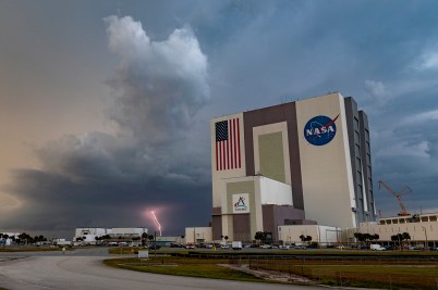 NASA's Vehicle Assembly Building at the Kennedy Space Center with an American flag and NASA logo visible on the exterior, set against a dramatic cloudy sky.