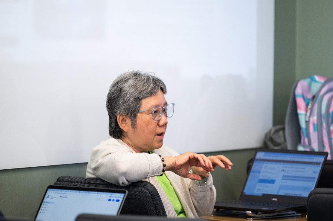 A teacher sitting in a chair in front of a laptop on a table talking.