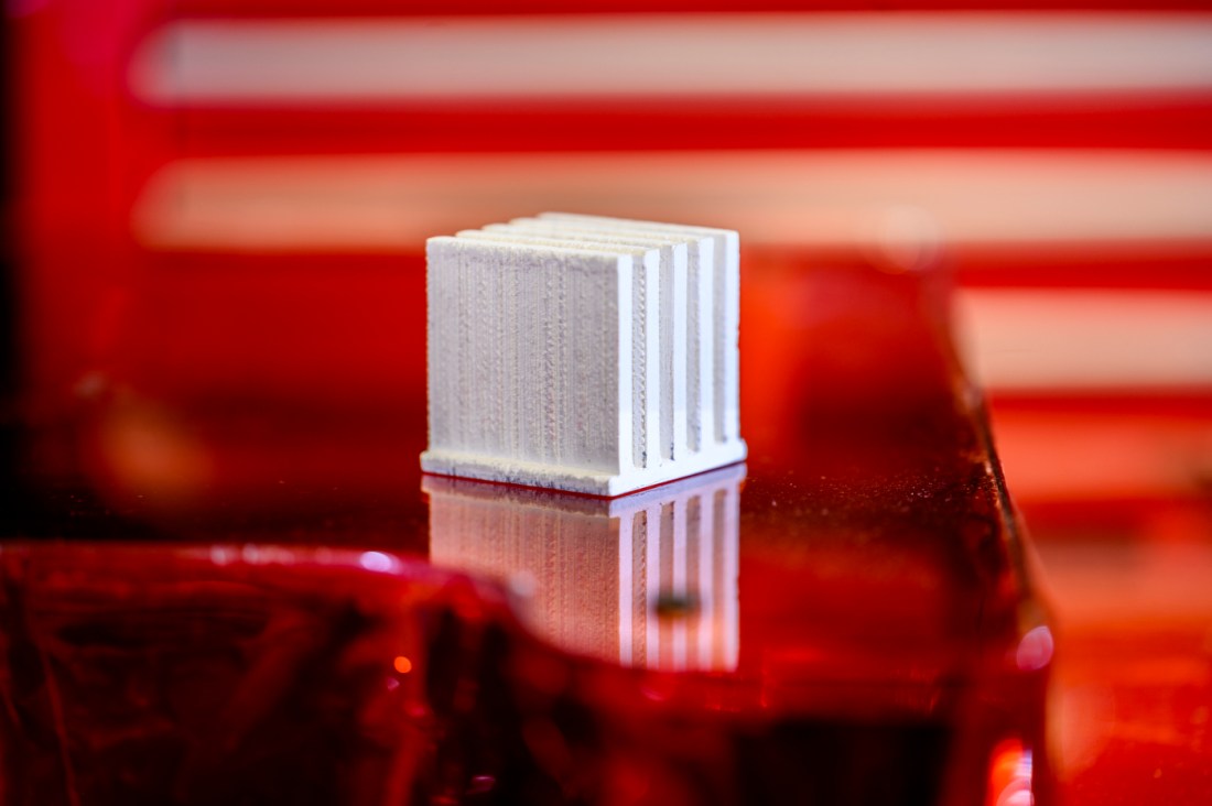 Small white cubic sample of 3D-printed heat-conductive plastic composite material with visible layered structure sitting on reflective red surface in laboratory setting.