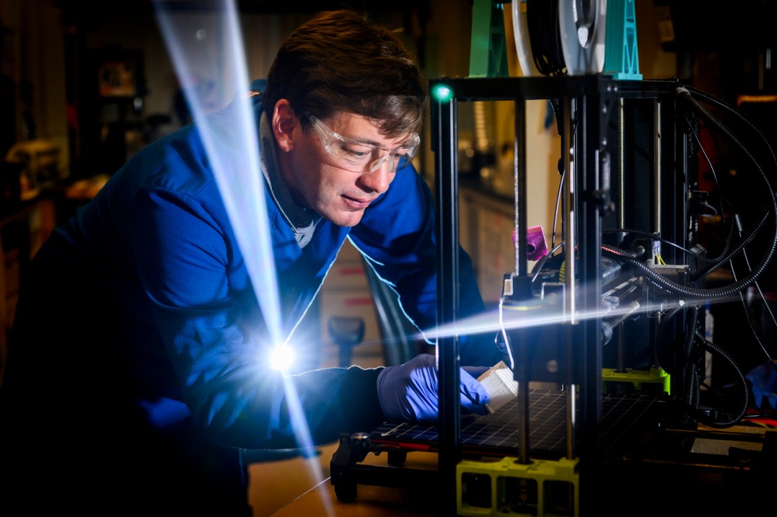 Randall Erb in blue shirt working with 3D printing equipment that emits bright white light in the Egan Research Center laboratory
