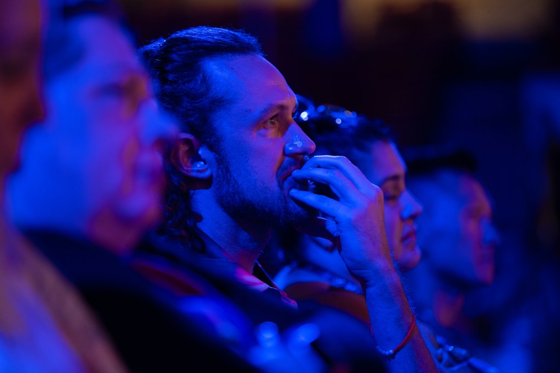 An audience member listens intently with their hand held up against their mouth in concentration. They are lit by blue and purple light.