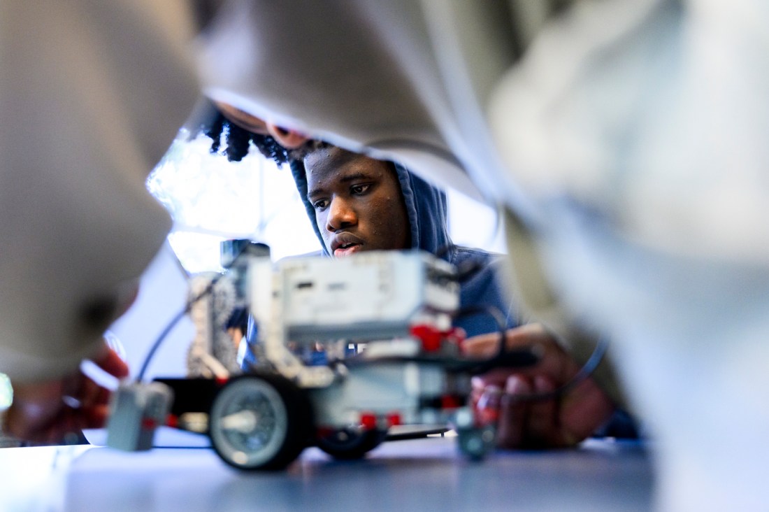 In the foreground a student leans over their robot making adjustments, in the background another student works on a separate robot.