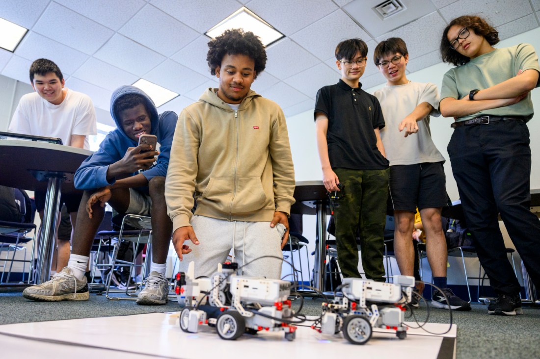 A student kneels in front of two lego robots, making final adjustments. Other students watch nearby, one is recording on their phone.