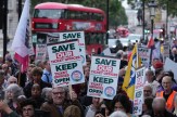 Protestors on strike in Britain holding picket signs. In the background, a red double decker bus drives on the street.