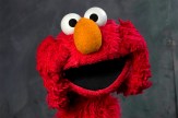 Elmo, a red furry Muppet character from Sesame Street, with his characteristic wide smile, googly eyes, and orange nose. He is up against a dark background.