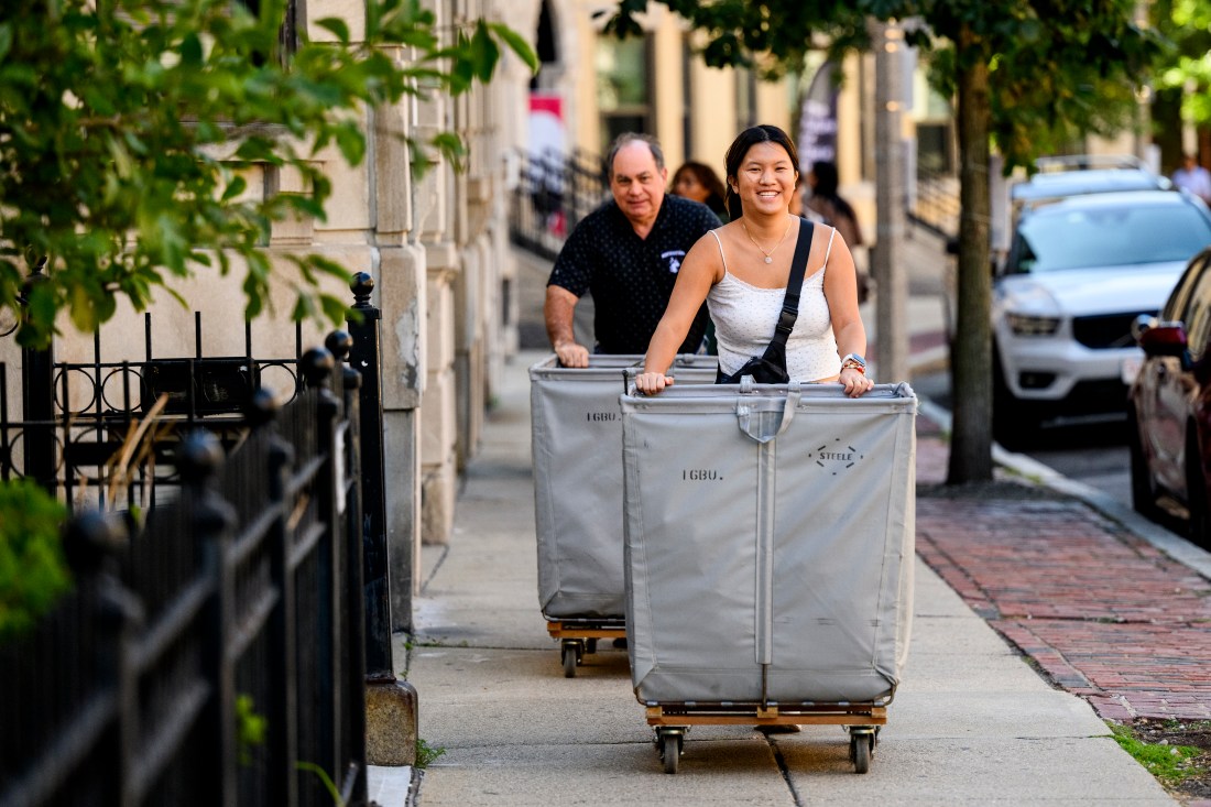 Two people push grey wheeled carts down the sidewalk. The one in front is smiling at the camera and wearing a white tank top. 