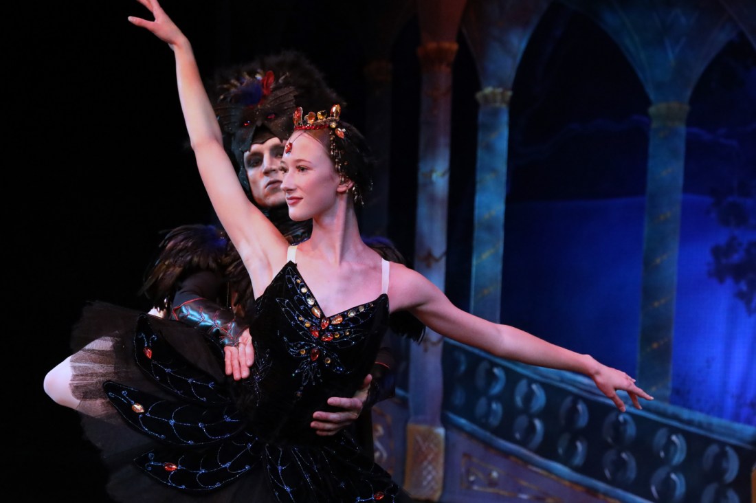 Two ballet dancers perform a pas de deux against a dark stage. The female dancer in a black tutu with red accents extends her arms in an elegant pose while her male partner in dark attire supports her. Blue-lit architectural elements and a decorative balustrade are visible in the background.