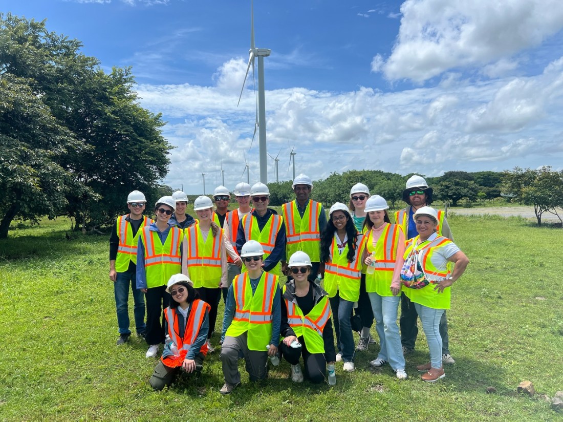 A group of Northeastern students wearing white hard hats and bright yellow and orange high-visibility safety vests post together on a grassy field. In the background are several wind turbines. It is a sunny day with clouds in the sky. 