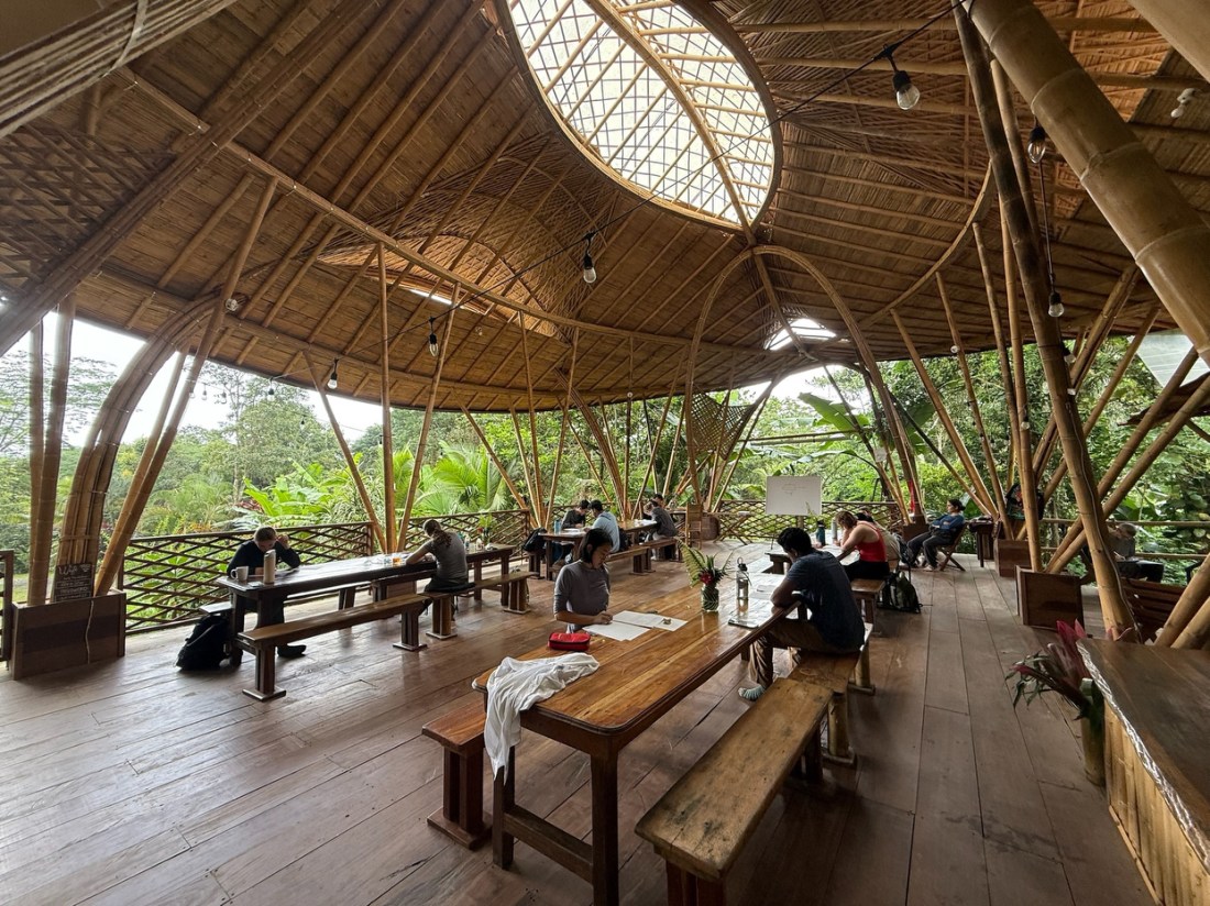 A bamboo pavilion with a curved architectural design and wooden picnic tables were people are engaged in their studies. 
