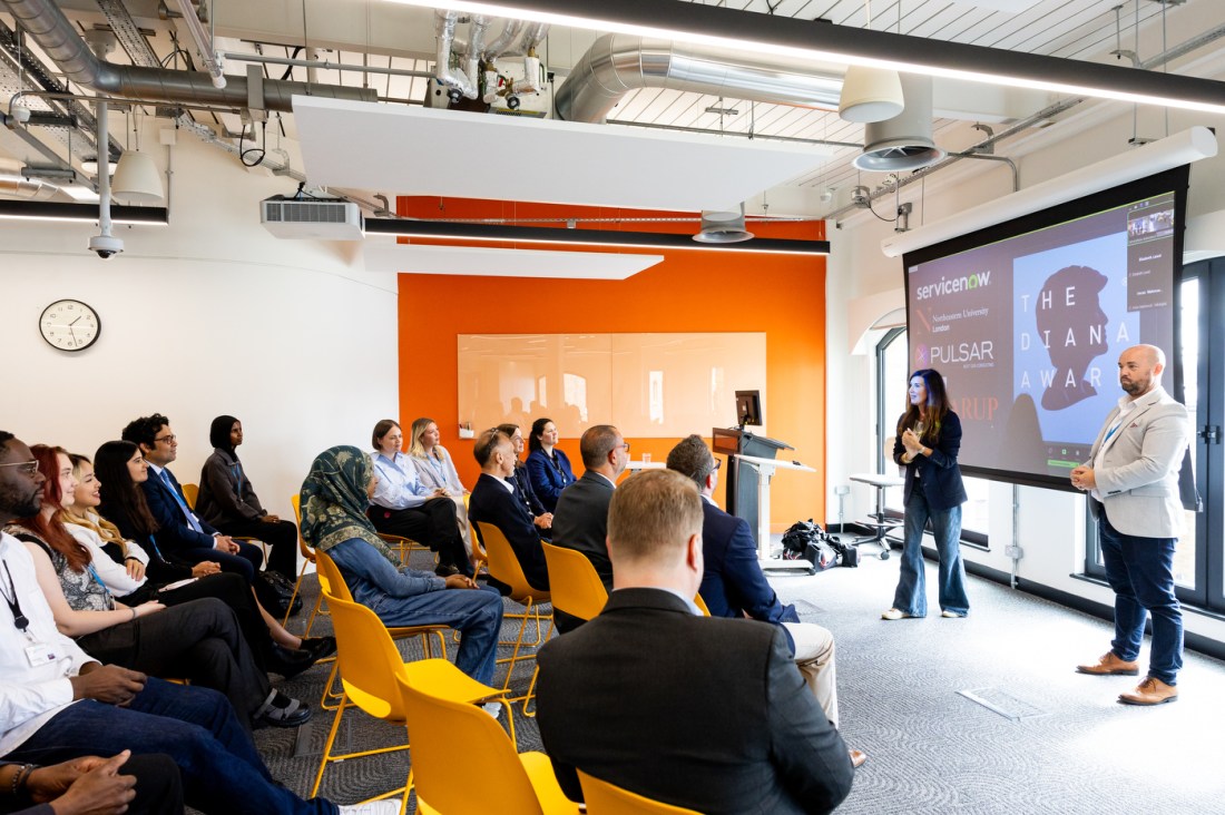 Audience members, including diverse students and guests, seated in yellow chairs attentively watching a graduation presentation in a modern classroom at Northeastern University’s London campus. An orange wall and digital screen are visible in the background.