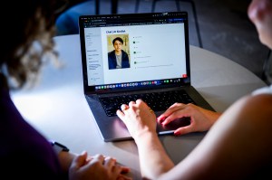 Two people sit at a table looking at a laptop screen that shows an AI chatbot interface with a profile photo of a person and text conversation.
