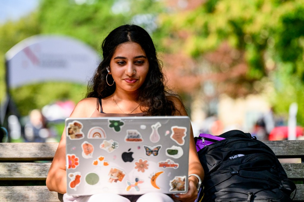 A student sits on a bench while working on a laptop, which is covered in stickers.