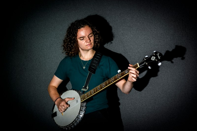 Maggie Heaney standing in front of a dark background playing the banjo.