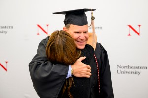 A Northeastern graduate in cap and gown smiles while hugging his wife during a small ceremony, celebrating the completion of a degree he began in the 1980s.