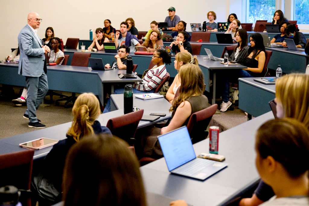 President Aoun in a suit stands at the front of a tiered classroom, speaking to rows of students seated at desks with laptops and water bottles.