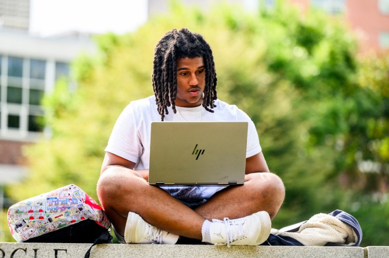 A person sits on a stone wall while working on a laptop on the Boston campus.