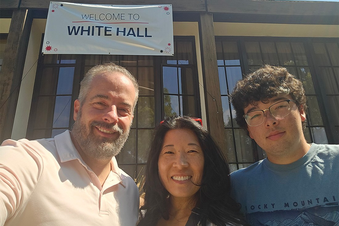 Sarah and Scott Pelletier taking a selfie in front of the White Hall building with their son Braden Pelletier. The building has a white banner that says 'Welcome to White Hall' hanging from the roof. 