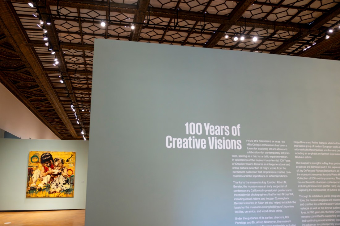 A museum gallery interior with a large wall display titled '100 Years of Creative Visions' with explanatory text. Behind it is another gallery well with a painting with many yellow tones. 