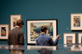 A Northeastern student and teacher observe various framed artworks hanging on a gallery wall of Mills College Art Museum.