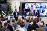People sitting in chairs on stage as part of a panel discussion in a modern classroom space at the Roux Institute. Behind them is a screen with images and the text &#039;Building the Future Together&#039; displayed.