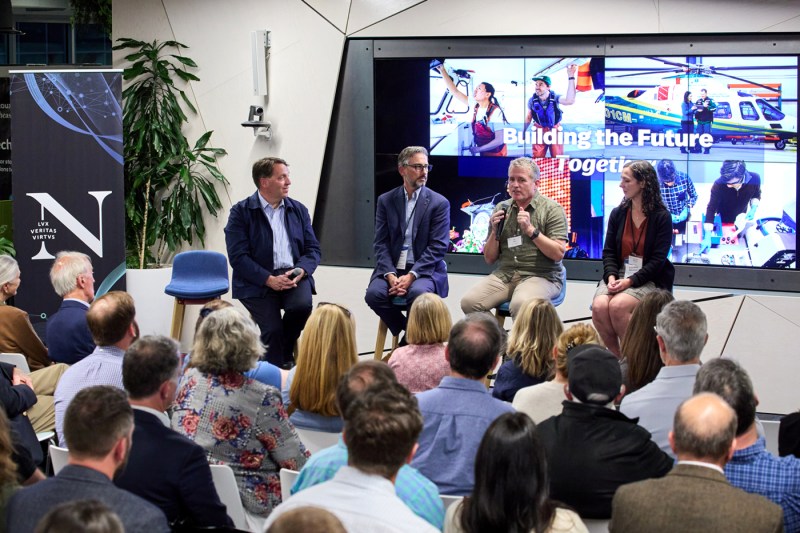 People sitting in chairs on stage as part of a panel discussion in a modern classroom space at the Roux Institute. Behind them is a screen with images and the text 'Building the Future Together' displayed.