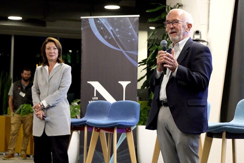 Two people standing on stage, one holding a microphone and speaking at the Roux Institute. 