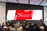 Northeastern University President Joseph E. Aoun sits next to Hemant Taneja on a platform during a fireside chat, with a screen depicting the Northeastern University logo raised above them.