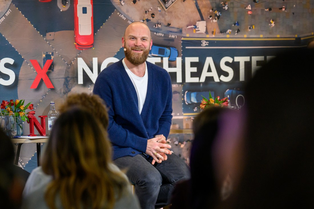 Coldplay drummer Will Champion sits on stage smiling while speaking with Northeastern students about AI and music at London’s Devon House.
