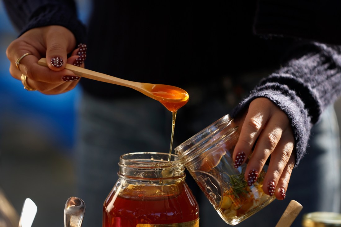 A person uses a wooden spoon to scoop honey out of a big jar into a smaller mason jar. 