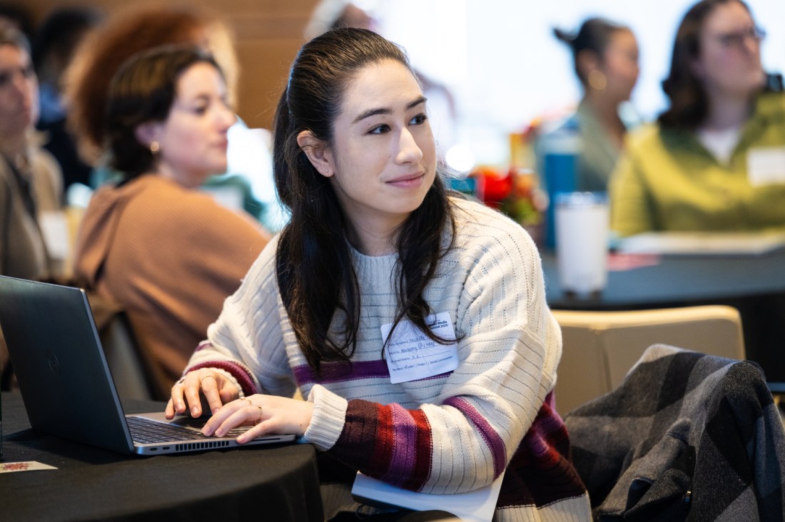 A person with long dark hair sitting at a table in front of a laptop during the Northeastern Social Media Summit. Other attendees are visible and blurred in the background. 