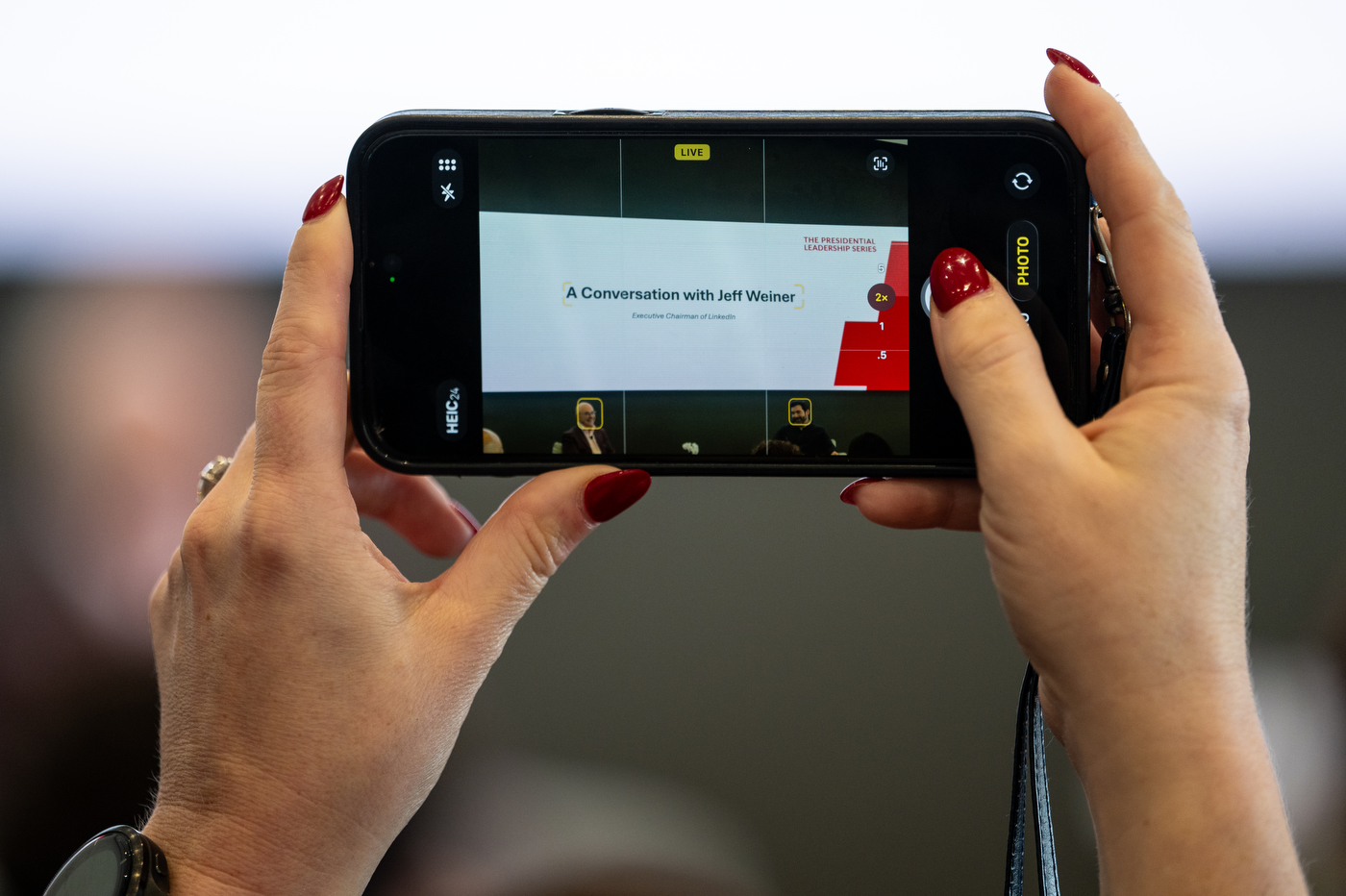 A person holds up a smartphone to photograph a presentation slide reading “A Conversation with Jeff Weiner” during a Northeastern University event.