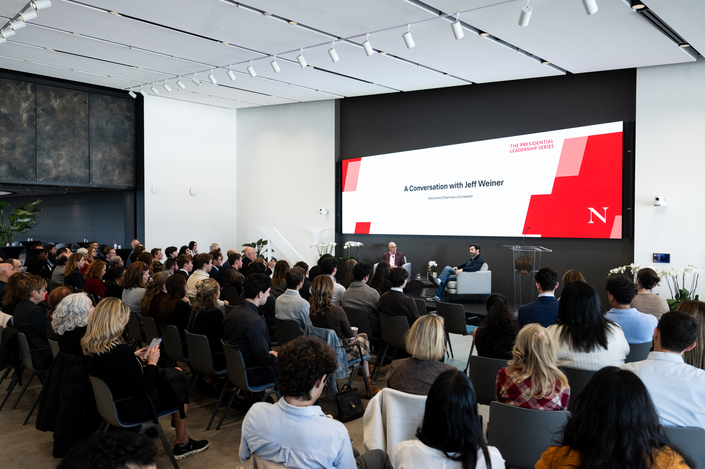 A large audience fills a modern auditorium at Northeastern University during a fireside chat between President Joseph E. Aoun and Jeff Weiner. A presentation screen displays the title “A Conversation with Jeff Weiner.”