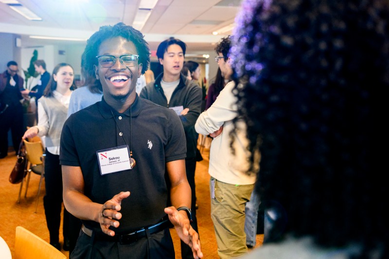 A Black student wearing a name tag that reads "Sekou" smiles while gesturing with both hands during the social event.