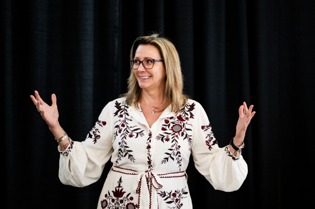 Illena Armstrong presents to an audience while gesturing with open hands. She is wearing a white embroidered dress and standing against a black curtain backdrop. 