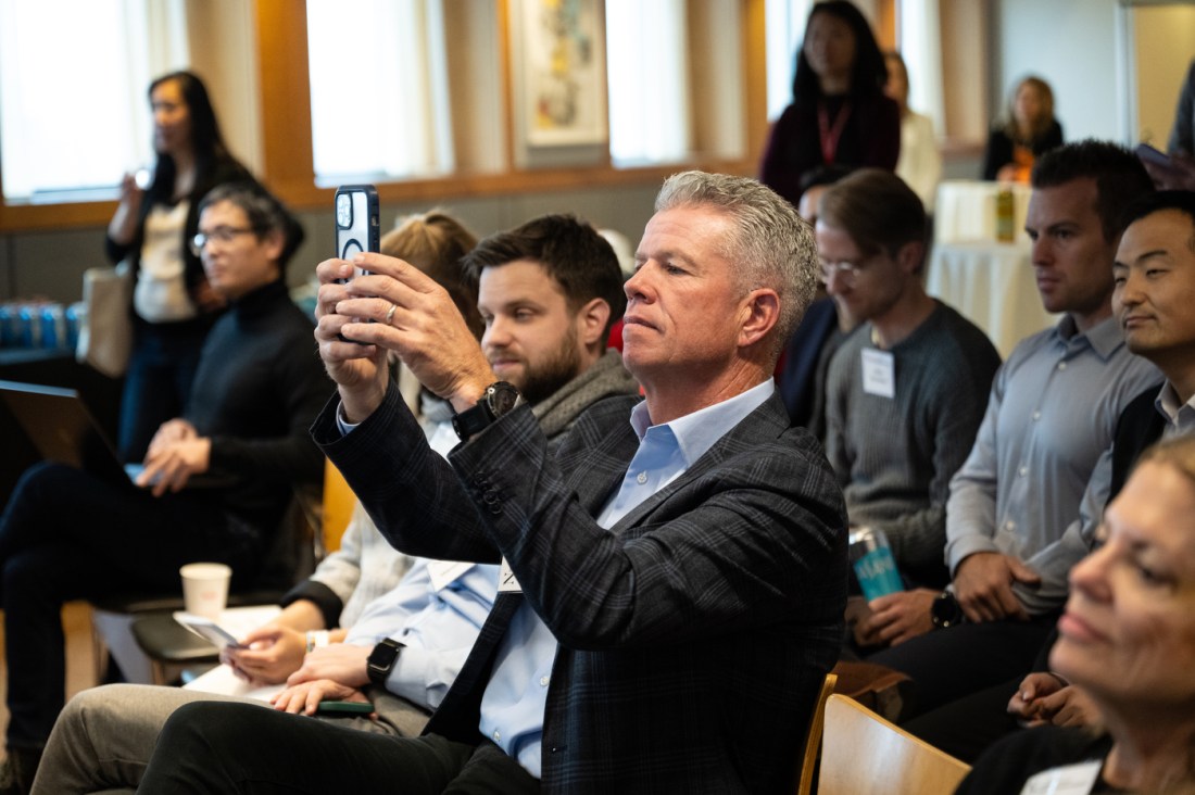 Audience members watch a presentation during DMSB Day at Northeastern University, with one attendee taking a photo using his phone.