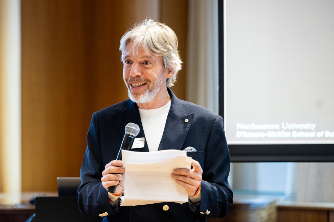 A speaker smiles while presenting with a microphone and notes in hand at DMSB Day in Northeastern’s Alumni Center.