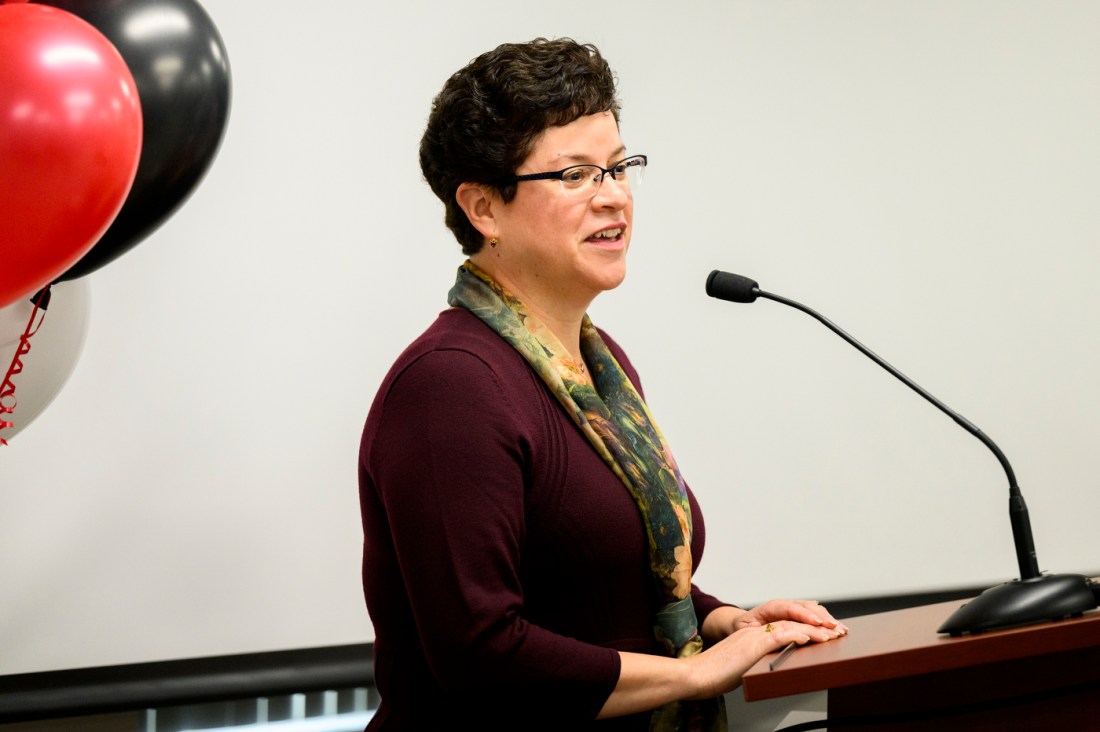 A woman with short dark hair and glasses speaks at a podium with a microphone. Red and black balloons are visible to the left side of the frame. 