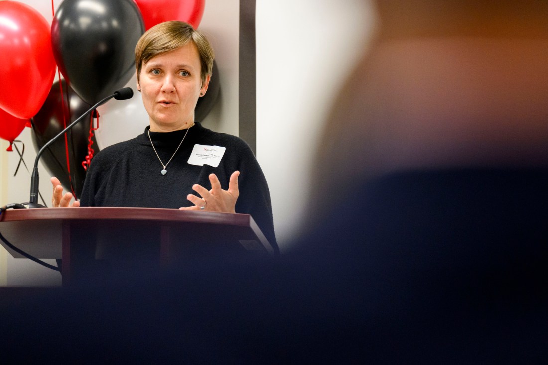 A woman with short hair wearing a dark shirt and a name tag stands in front of a podium with a microphone speaking at the Fall River campus grand opening. Behind her are red and black balloons. 