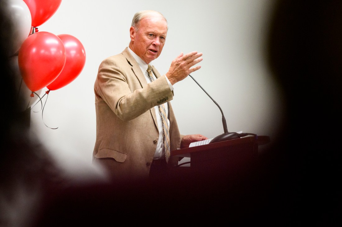 A man in a tan blazer gestures while speaking at a podium. Red and black balloons are visible behind him. 